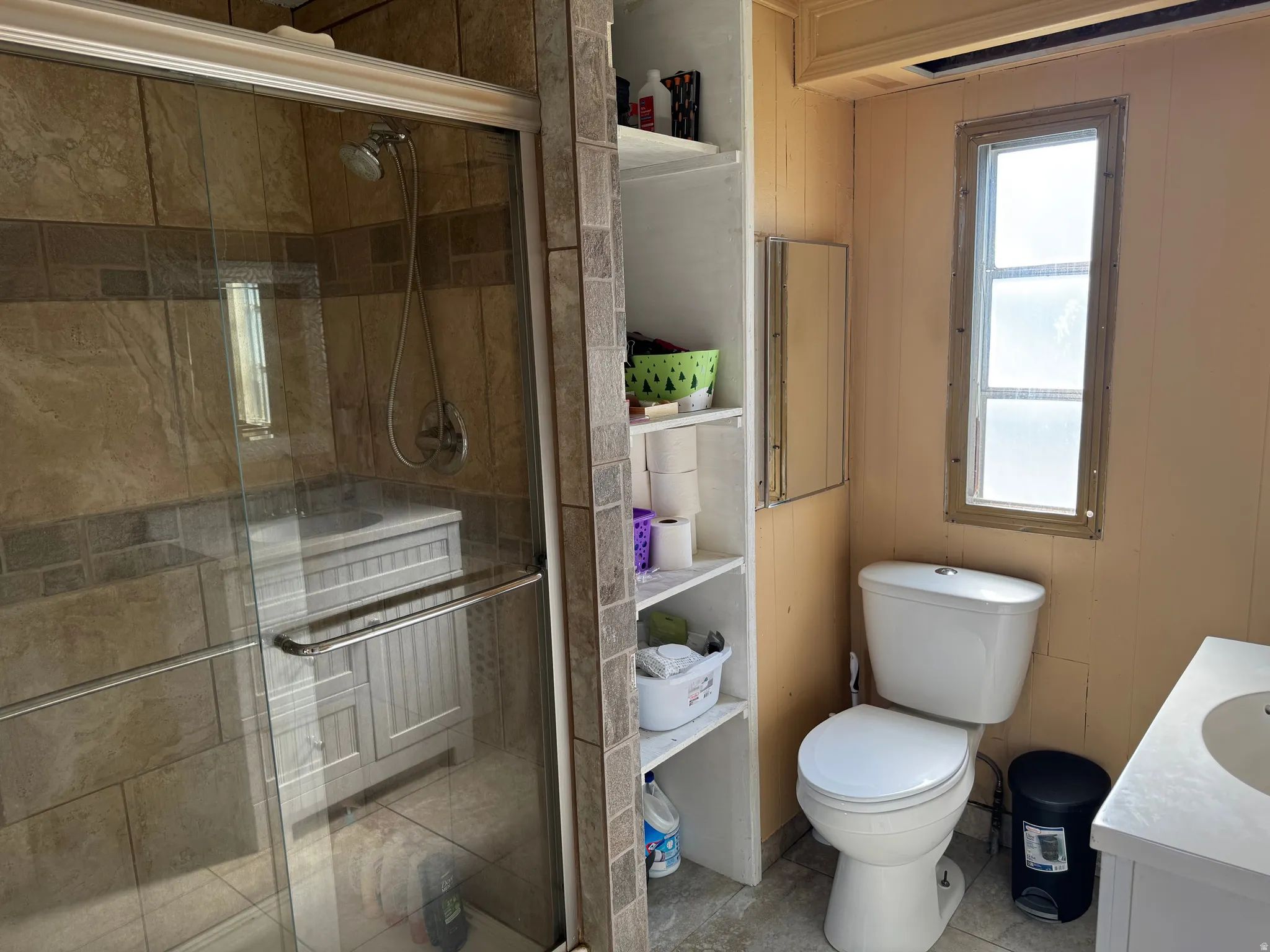 Bathroom featuring a tiled shower enclosure with glass sliding doors, built-in shelving, a single-pane window, and a white toilet