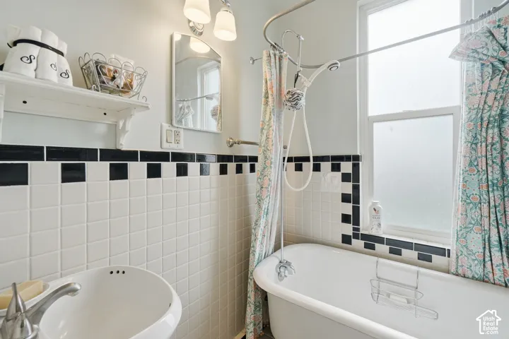 White tiled bathroom featuring a clawfoot tub with an overhead shower, a pedestal sink, and a frosted window