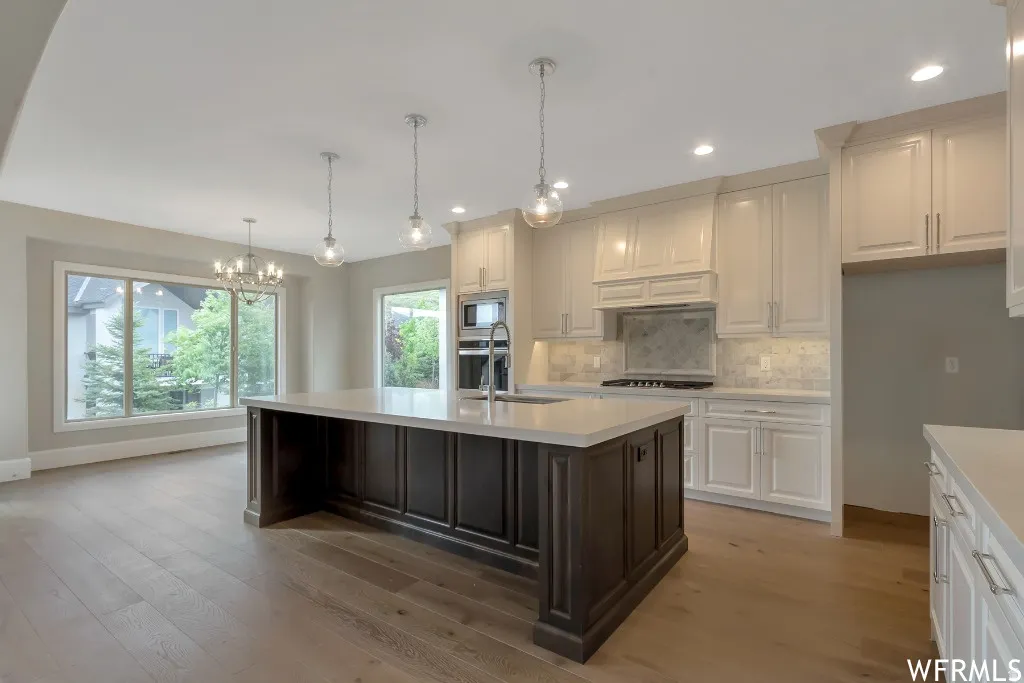 Kitchen with two tone color scheme, tasteful backsplash, suspended lighting, light wood-style flooring, and an island with sink