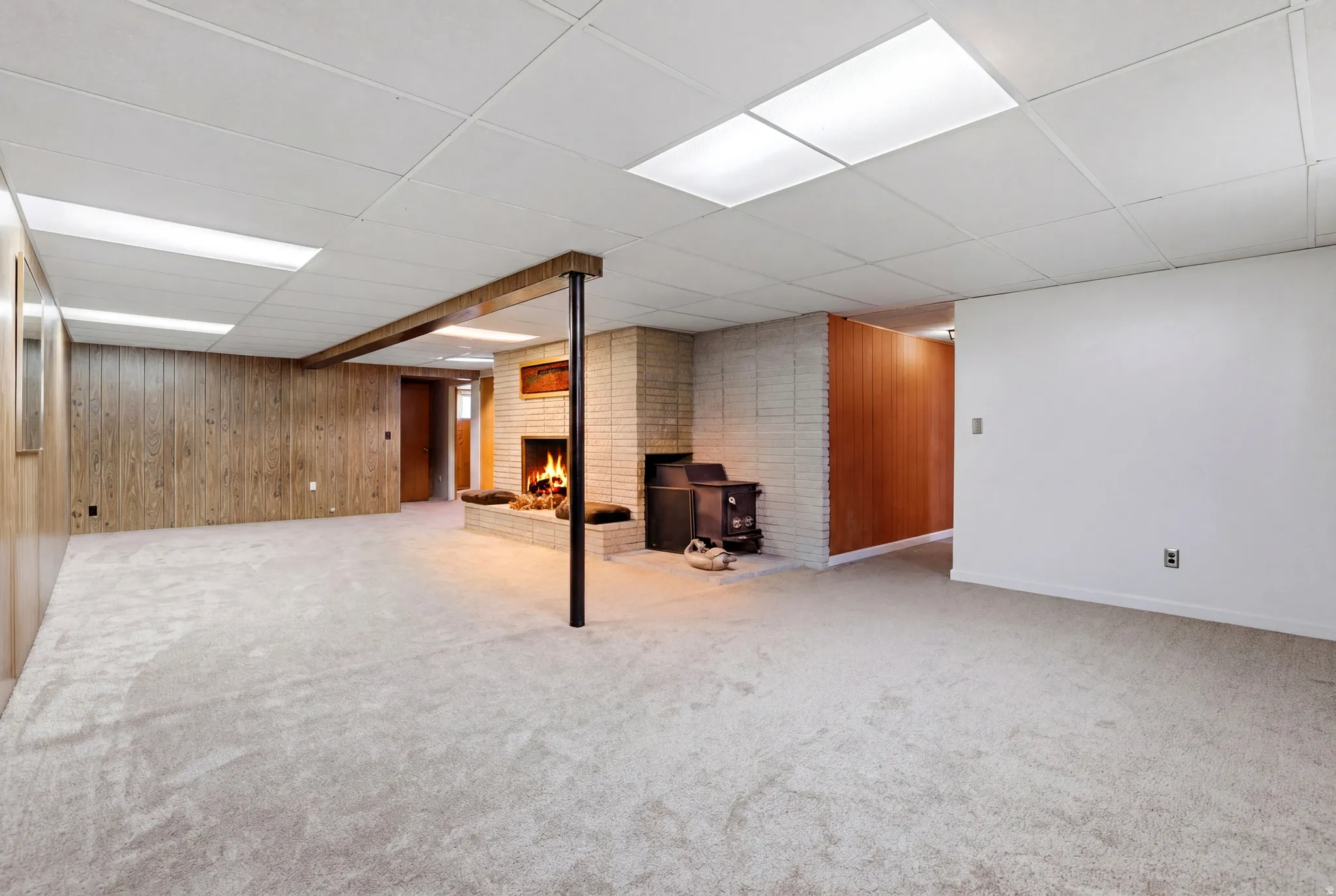 Finished basement featuring a brick fireplace, wooden walls, a drop ceiling, and light colored carpet