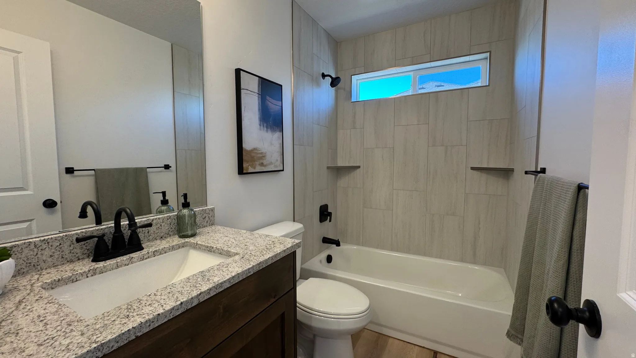 Bathroom featuring a granite countertop vanity with an undermount sink, dark wood-finish cabinetry, a large wall-mounted mirror, a tub/shower combination with tile surround, and a horizontal transom window