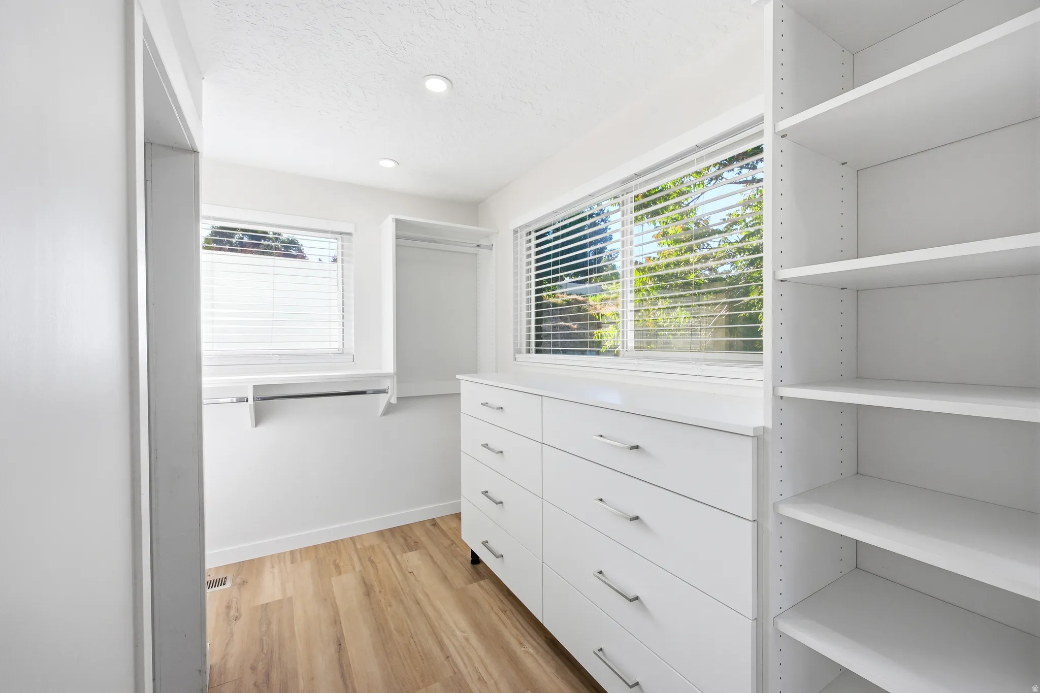 Custom closet system featuring wood-finish flooring, recessed lighting, built-in shelving, integrated drawers with metal hardware, and natural light from horizontal windows