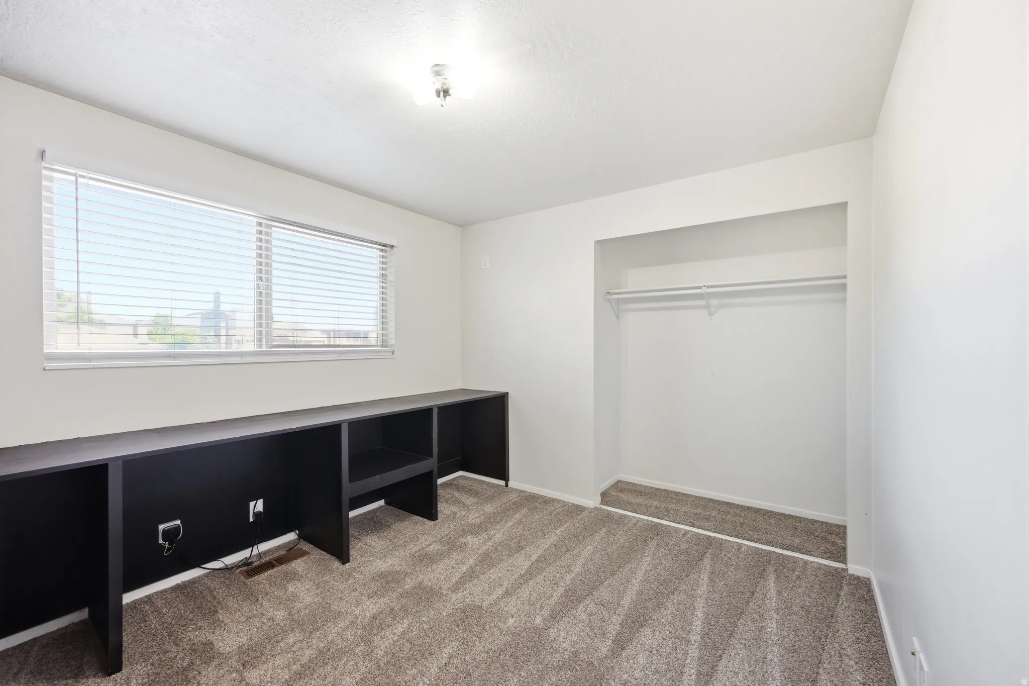 Room featuring a large window with blinds, a built-in dark storage and desk unit, wall-to-wall carpeting, and an open closet with a hanging rod