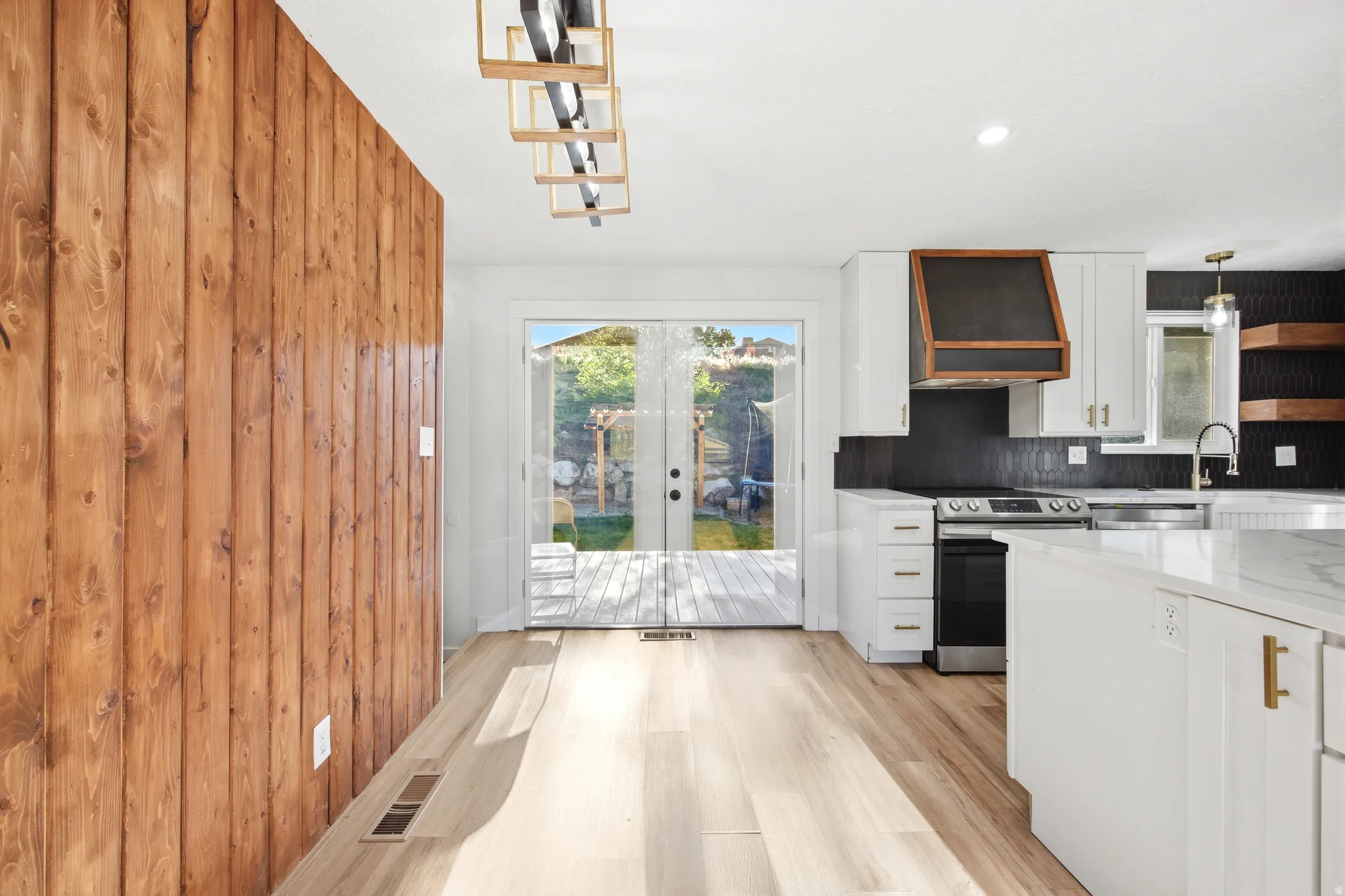 Open-concept kitchen and dining area featuring light wood-finish flooring, a wood-paneled accent wall, white cabinetry with gold hardware, a built-in range hood with wood trim, and double glass doors leading to an exterior deck