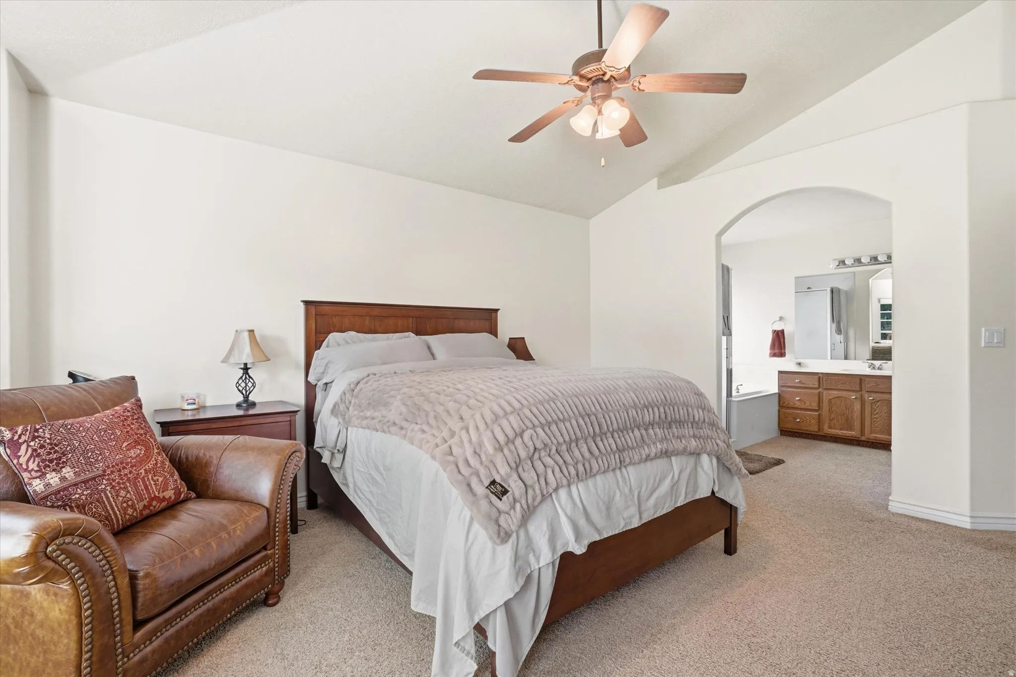 Spacious sleeping area with vaulted ceiling and ceiling fan, featuring an arched doorway leading to a private bath with a double vanity and built-in tub