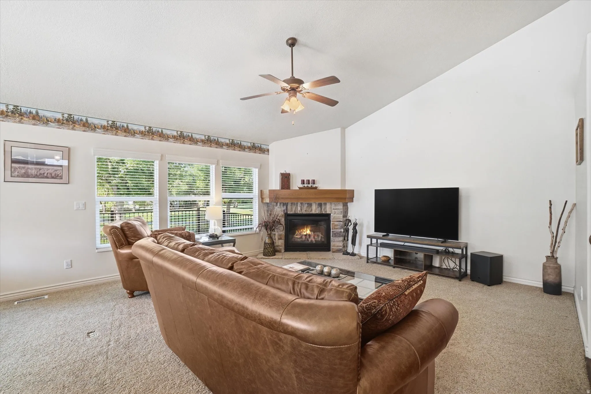 Living area featuring a corner stone fireplace with a wood mantle, vaulted ceiling, and ceiling fan