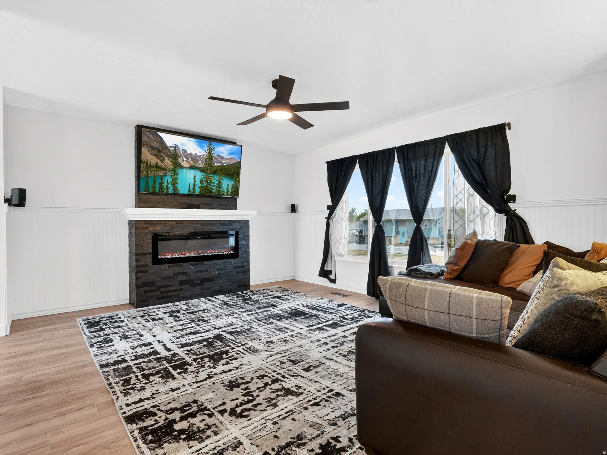 Living room with a wainscoted wall, light wood finished floors, a glass covered fireplace, and a ceiling fan