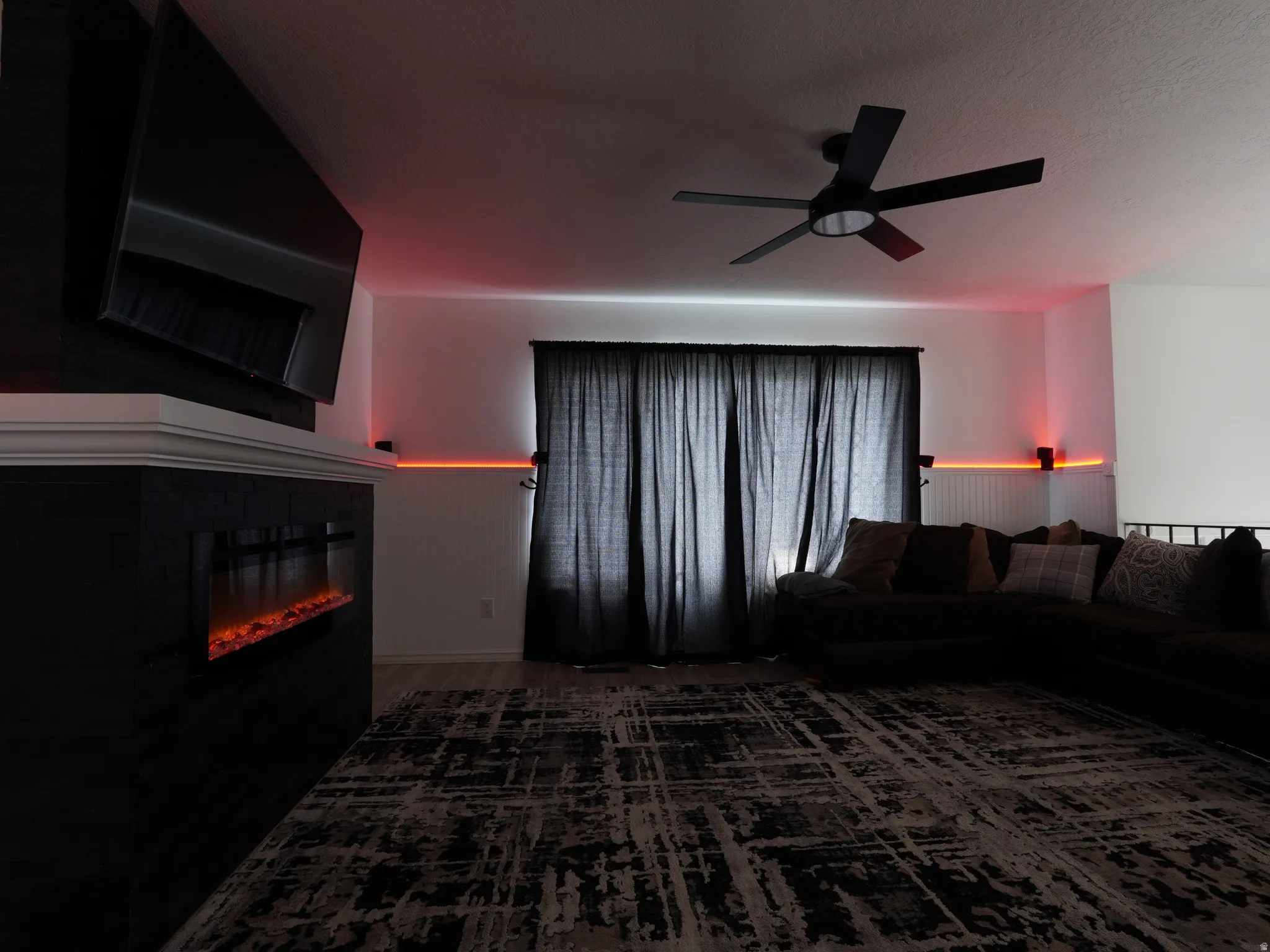 Living area featuring ceiling fan, a glass covered fireplace, wood finished floors, a textured ceiling, and wainscoting