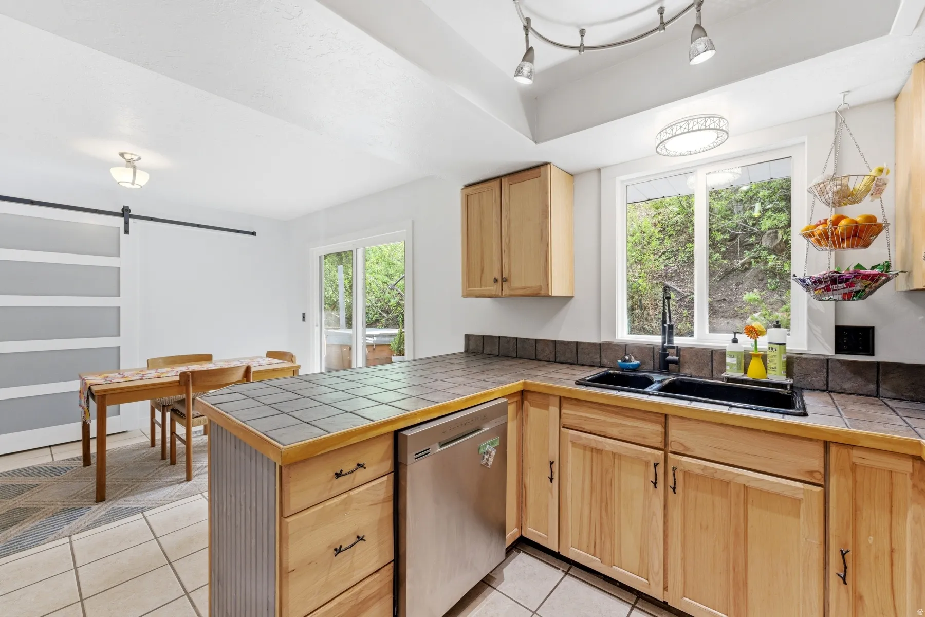 Kitchen featuring a barn door, light wood finish cabinetry, stainless steel dishwasher, light tile patterned floors, and a peninsula