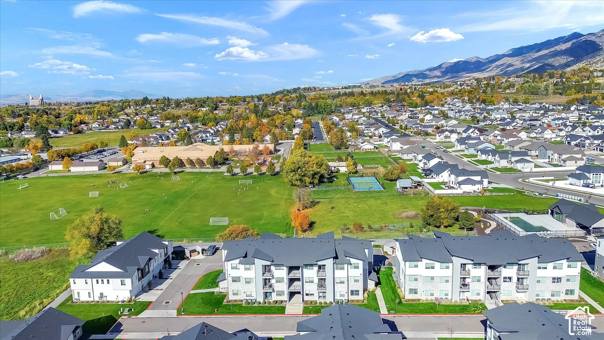 Aerial perspective of multi-family residences featuring dark-toned pitched roofs, light-colored exteriors, balcony spaces, and proximity to open green space with mountainous backdrops