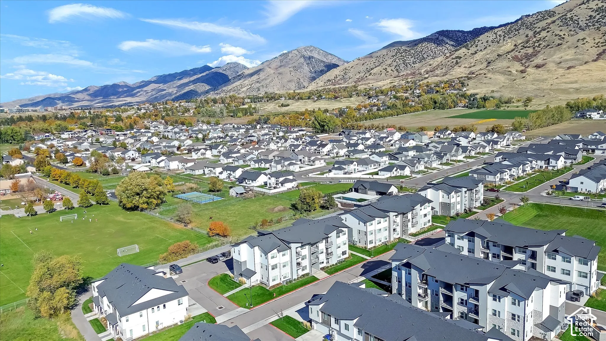 Aerial perspective of multi-family residences featuring dark-toned pitched roofs, light-colored exteriors, balcony structures, and proximity to expansive open green space with mountain backdrops