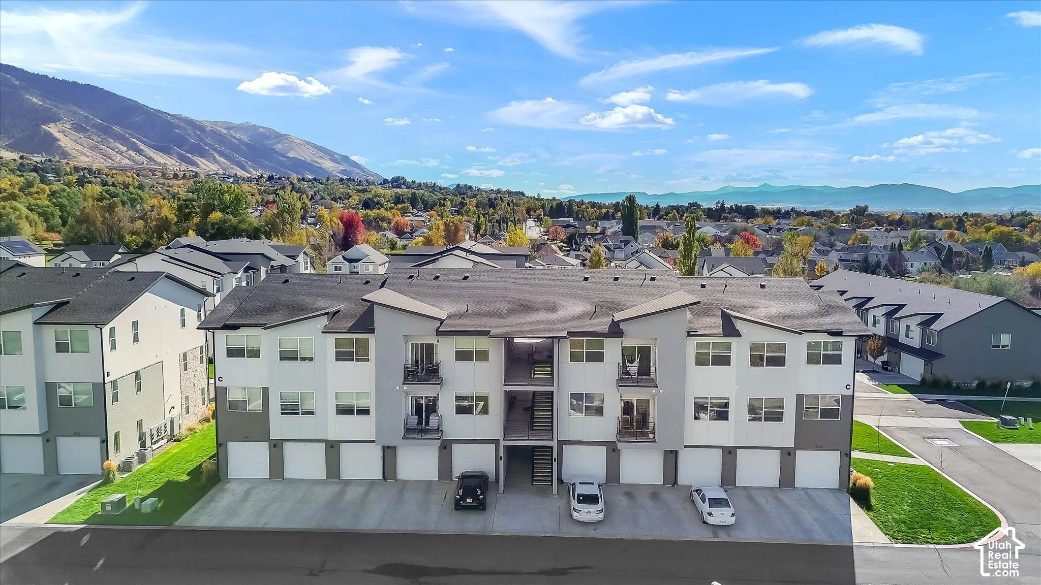 Multi-level residential building featuring a two-tone gray and white exterior, shingled roofing, attached ground-level garages, and private balconies, set against a prominent mountain backdrop