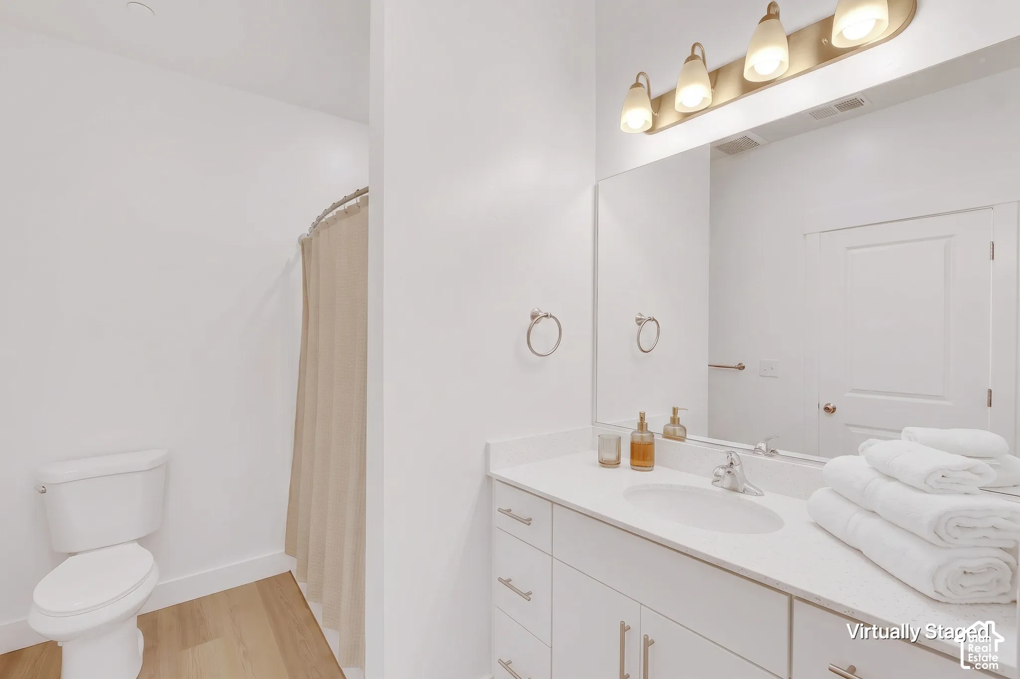 Bathroom featuring a vanity with a white countertop and integrated sink, brushed metal hardware, a wall-spanning mirror, overhead multi-bulb lighting, and wood-finish flooring