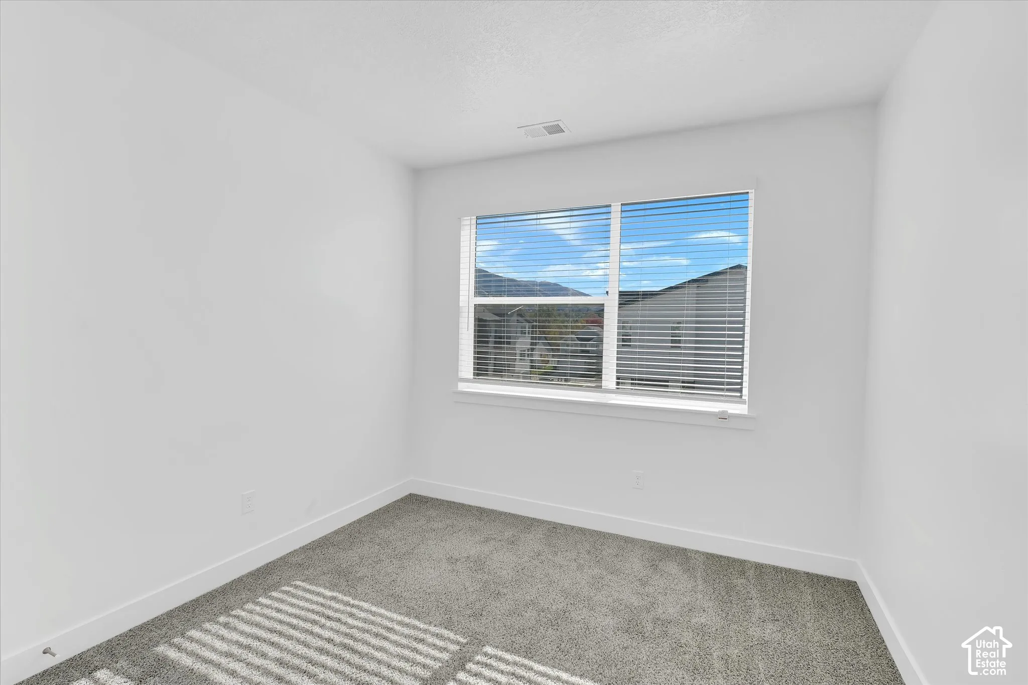 Neutral-toned interior room featuring gray wall-to-wall carpeting, crisp white walls, and a large horizontal sliding window with horizontal blinds