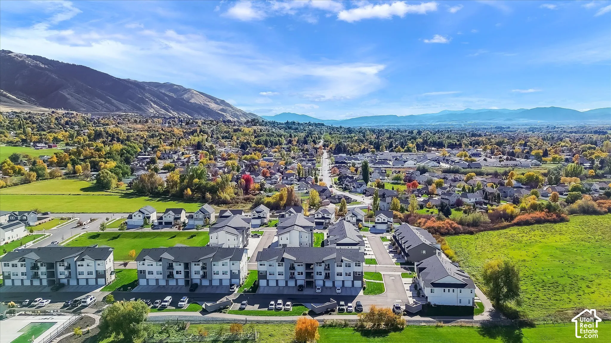 Aerial perspective of multi-family residences featuring dark-toned pitched roofs, light-colored siding, and paved parking lots, set against a backdrop of mountains and open green space