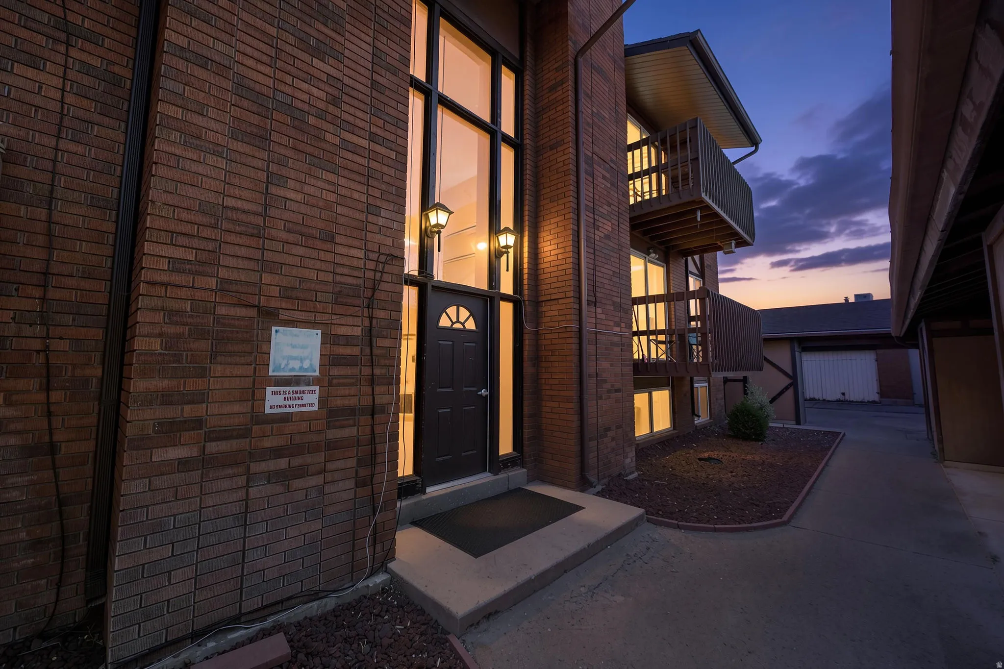 Brick facade building featuring a full-light entry door with an arched transom, exterior wall sconces, multi-level balconies with wood railings, and a concrete walkway