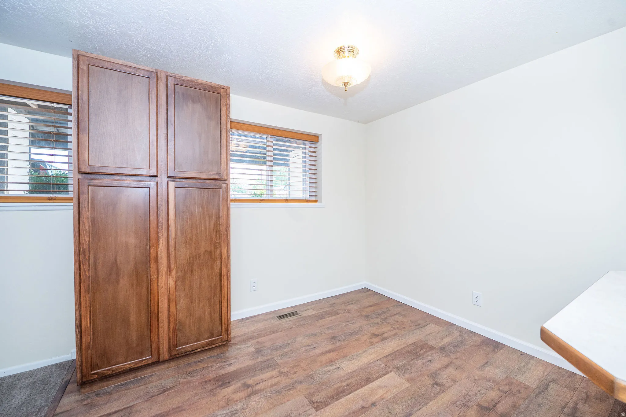 Hardwood-style flooring with a built-in wood-finish storage cabinet, two windows with blinds, and white walls
