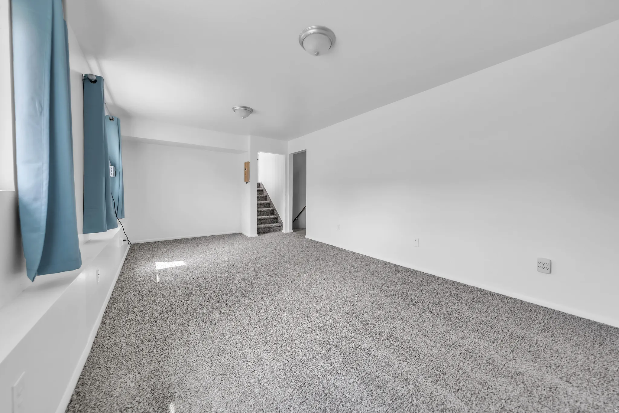 Spacious room featuring light gray carpeting, white walls, two flush-mount ceiling lights, and a visible staircase leading to an upper level