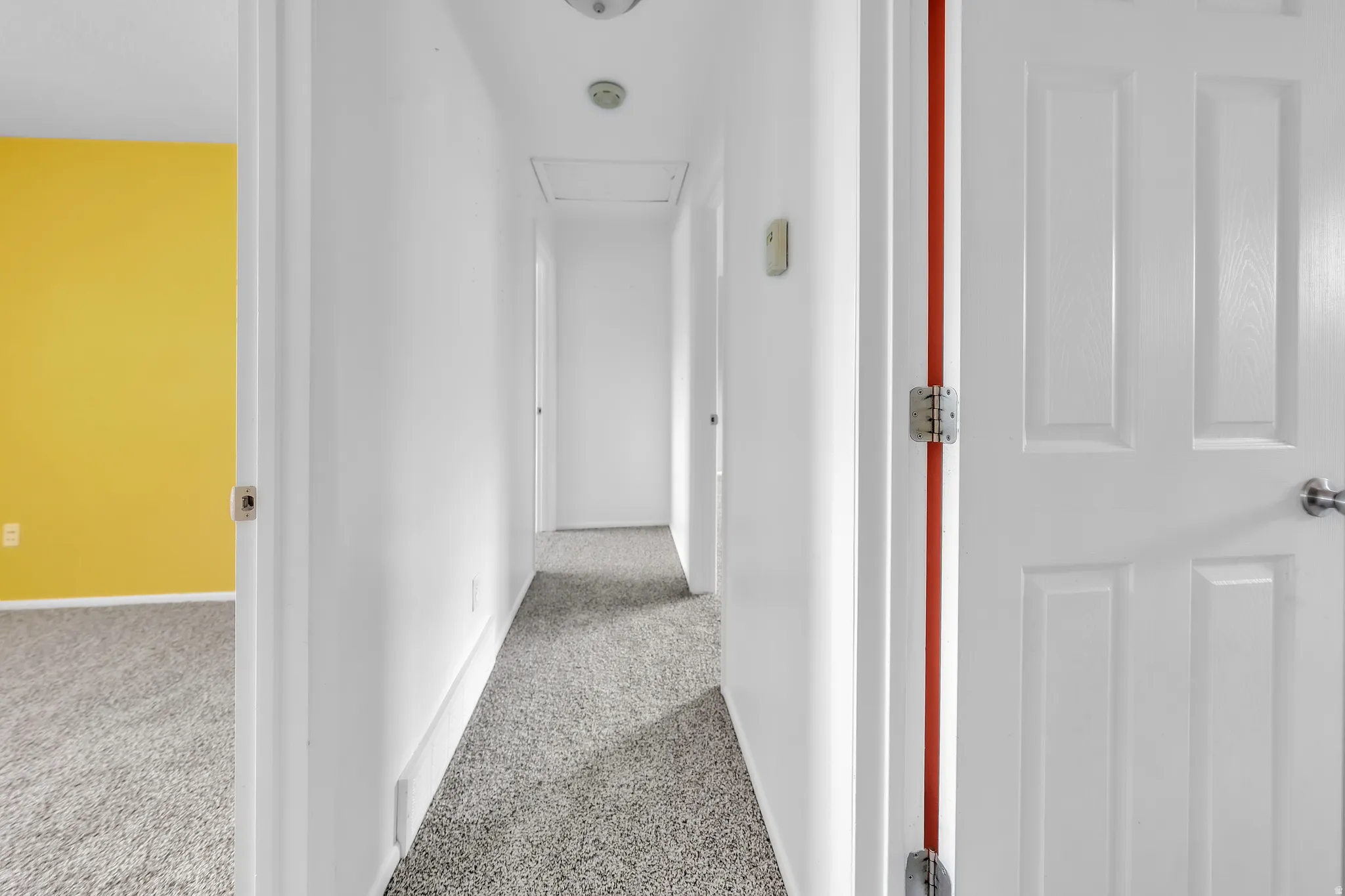 Carpeted hallway with white painted walls and trim, featuring overhead flush-mount lighting and a panel door with satin nickel hardware
