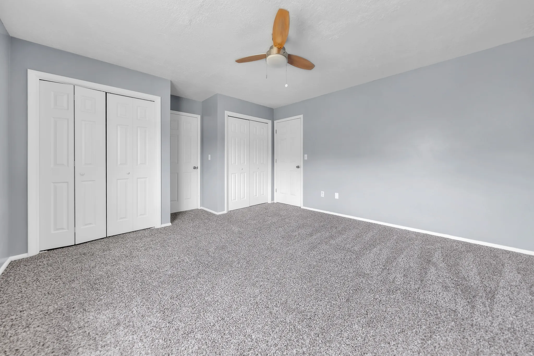 Spacious room featuring light gray wall paint, gray carpet flooring, and a ceiling fan with wooden blades