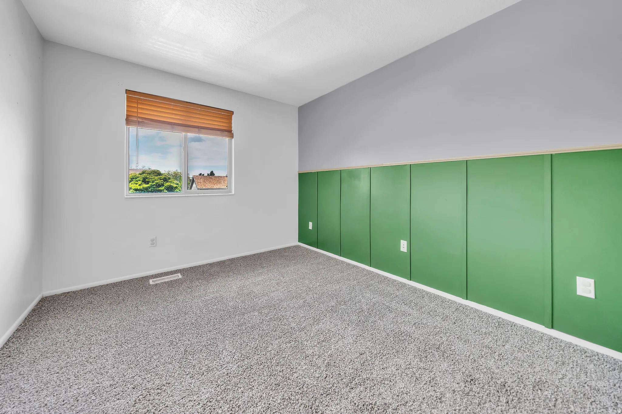 Room featuring a large window with a wood-finish blind, textured ceiling, light gray walls, and a green accent panel wall with white trim