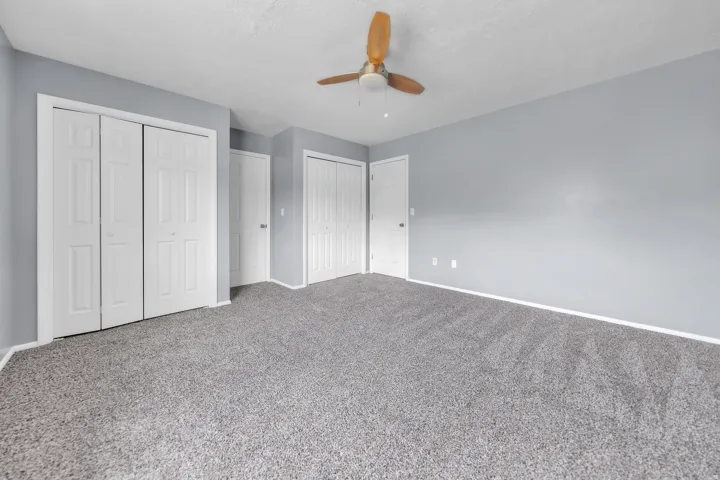 Spacious room featuring light gray wall paint, gray carpet flooring, and a ceiling fan with wooden blades