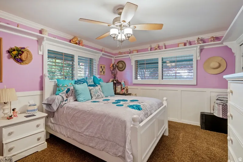 Bedroom with crown molding, a wainscoted wall, dark colored carpet, ceiling fan, and a decorative wall