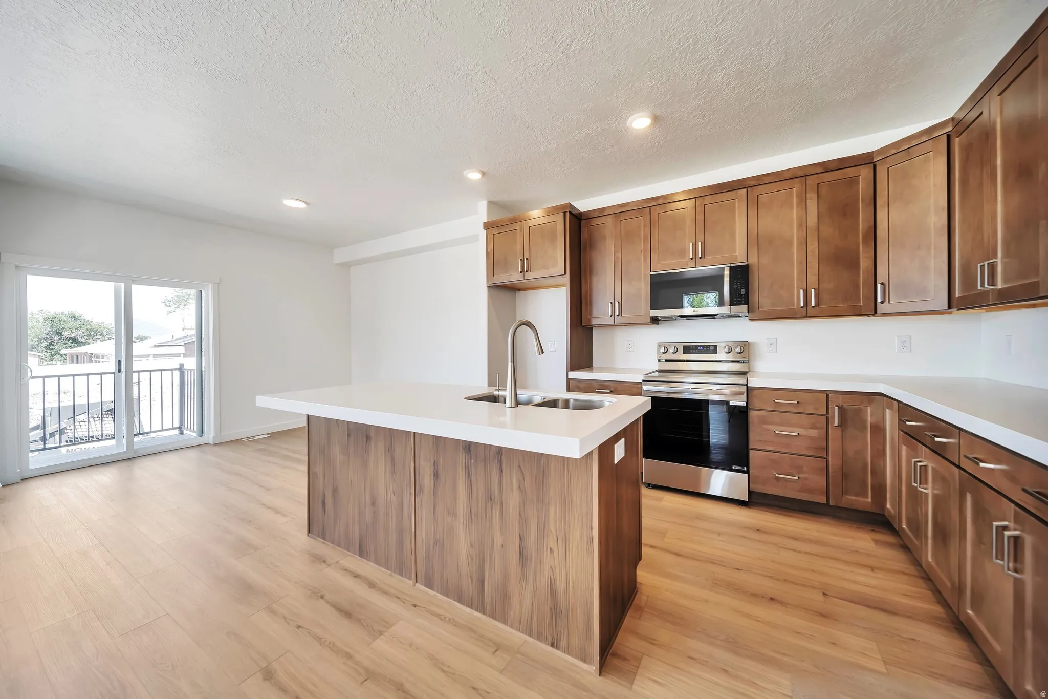 Kitchen featuring stainless steel appliances, a center island with sink, wood finish cabinets, light countertops, and recessed lighting