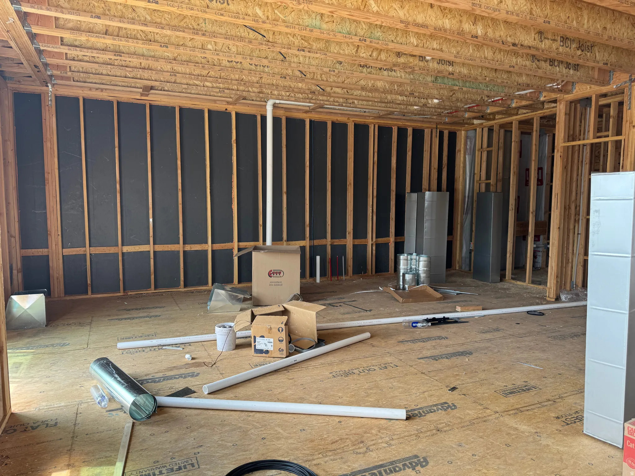 Wood stud framing in an unfinished interior space, featuring a plywood subfloor, exposed ceiling joists, and black insulation