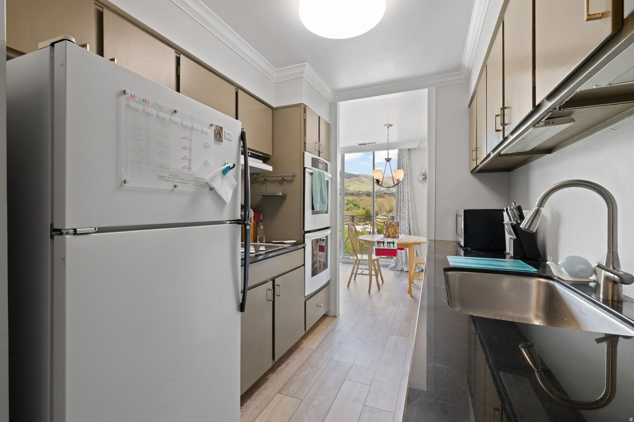 Kitchen with white appliances, ornamental molding, hanging light fixtures, dark countertops, and floor to ceiling windows