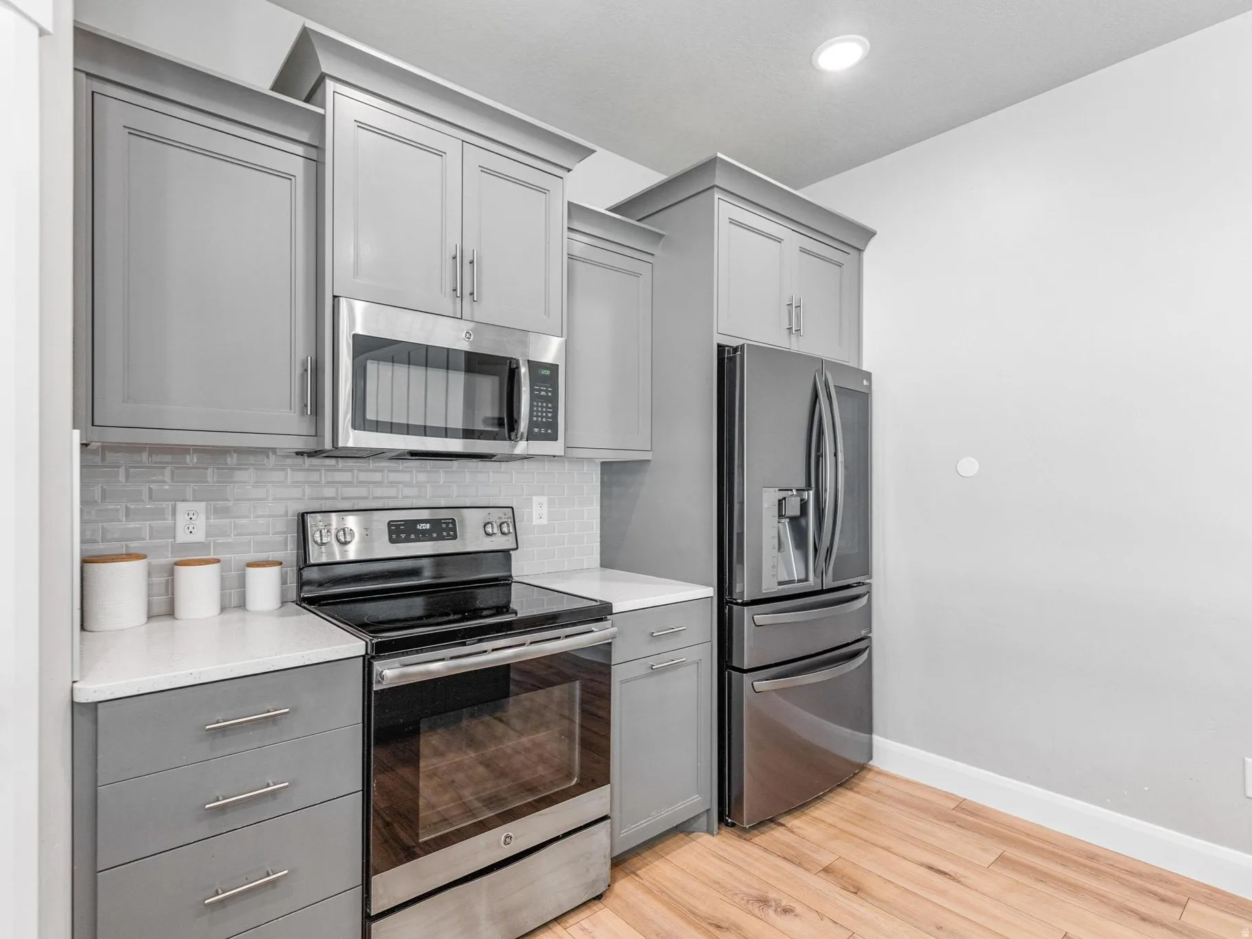 Kitchen with gray cabinetry, stainless steel appliances, light wood-type flooring, decorative backsplash, and light stone counters