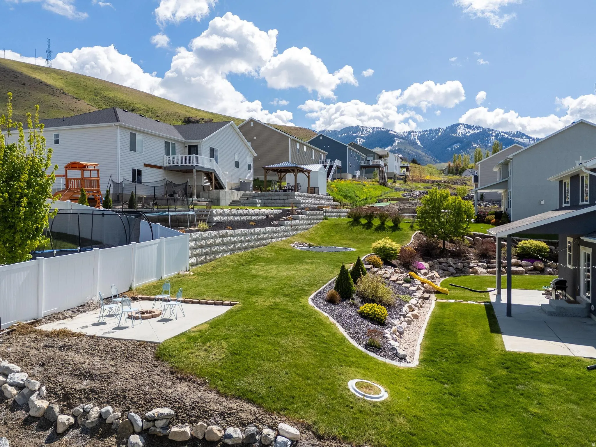 Tiered landscaping with retaining walls, established plantings, covered patio, and a dedicated fire pit area