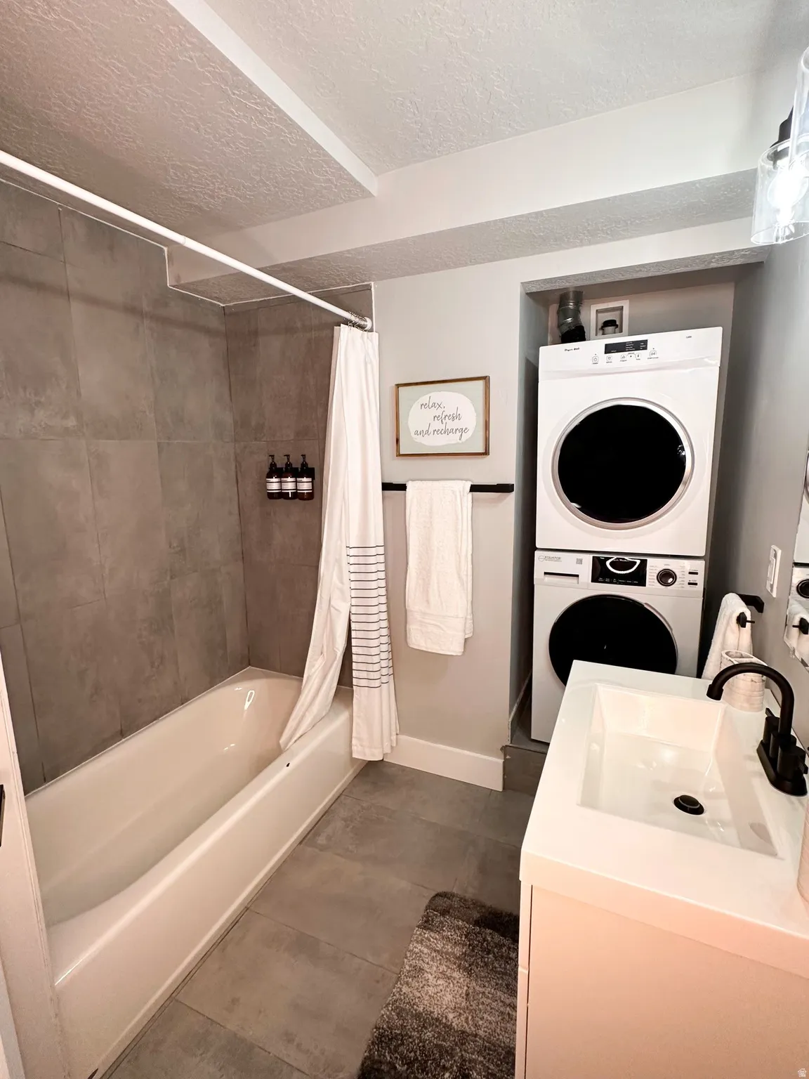 Furnished Unit #44 Full bath featuring vanity, stacked washer / drying machine, a textured ceiling, shower / bath combination with curtain, and dark tile patterned floors