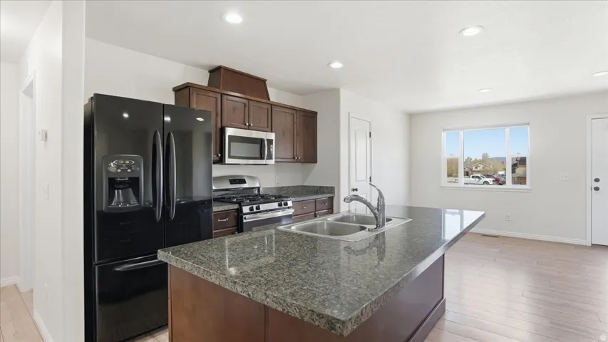Kitchen featuring stainless steel appliances, dark wood finish cabinetry, light wood-type flooring, dark stone counters, and a center island with sink