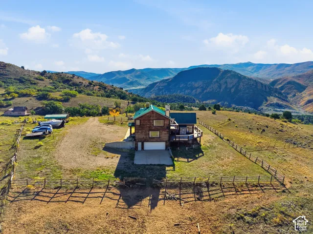 Two-story wood-sided residence featuring a metal roof, wraparound deck, attached two-car garage, and perimeter fencing on a multi-acre parcel set against a mountain backdrop