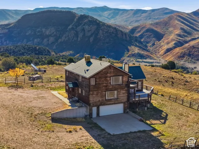 Two-story home with wood siding, a green metal roof, a concrete driveway, a two-car garage, and an elevated wood deck, set against a backdrop of mountainous terrain