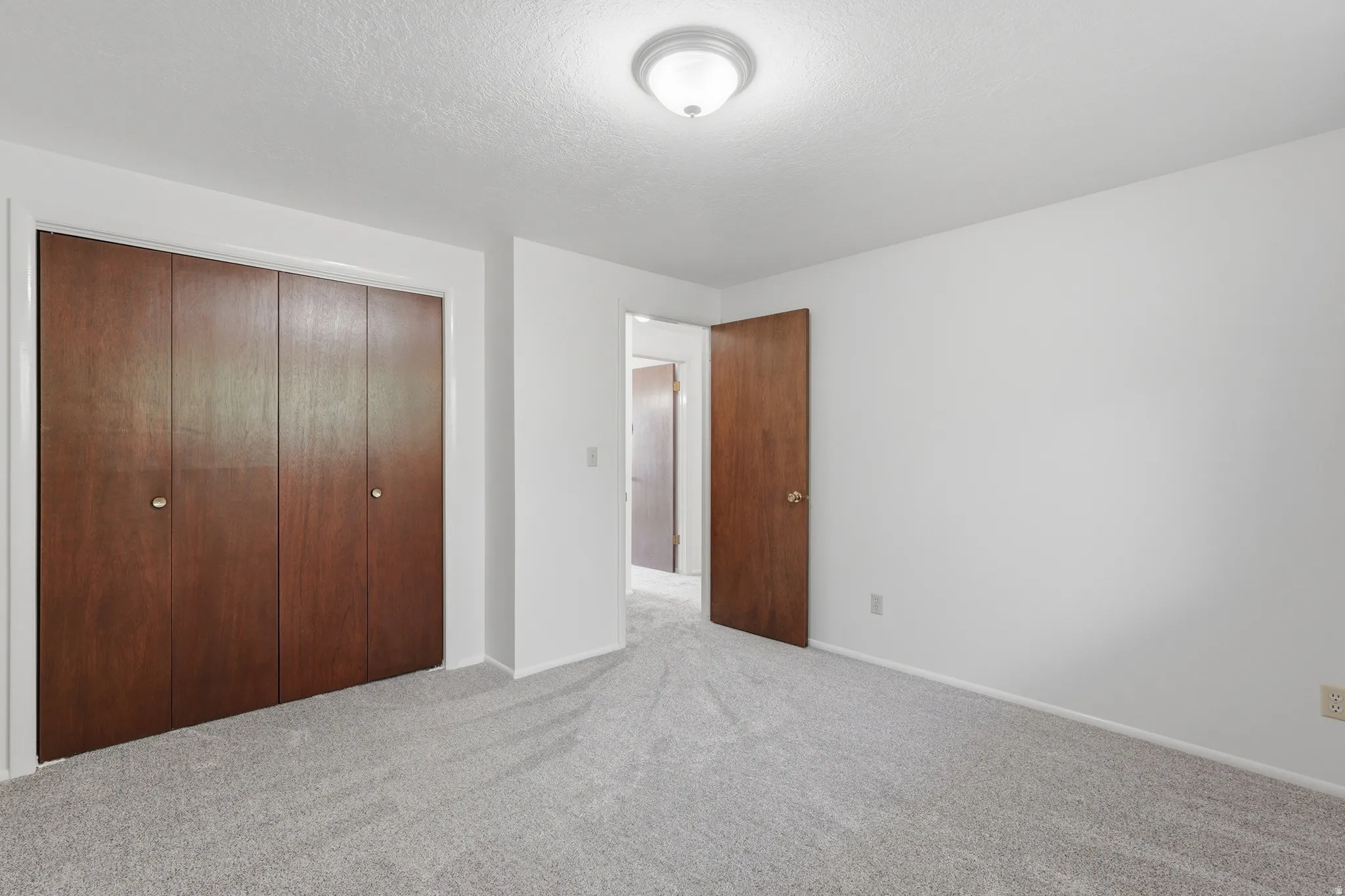 Neutral paint tones and light-grey carpeting define this interior room, featuring wood-finish bi-fold closet doors and a flush-mount ceiling light fixture