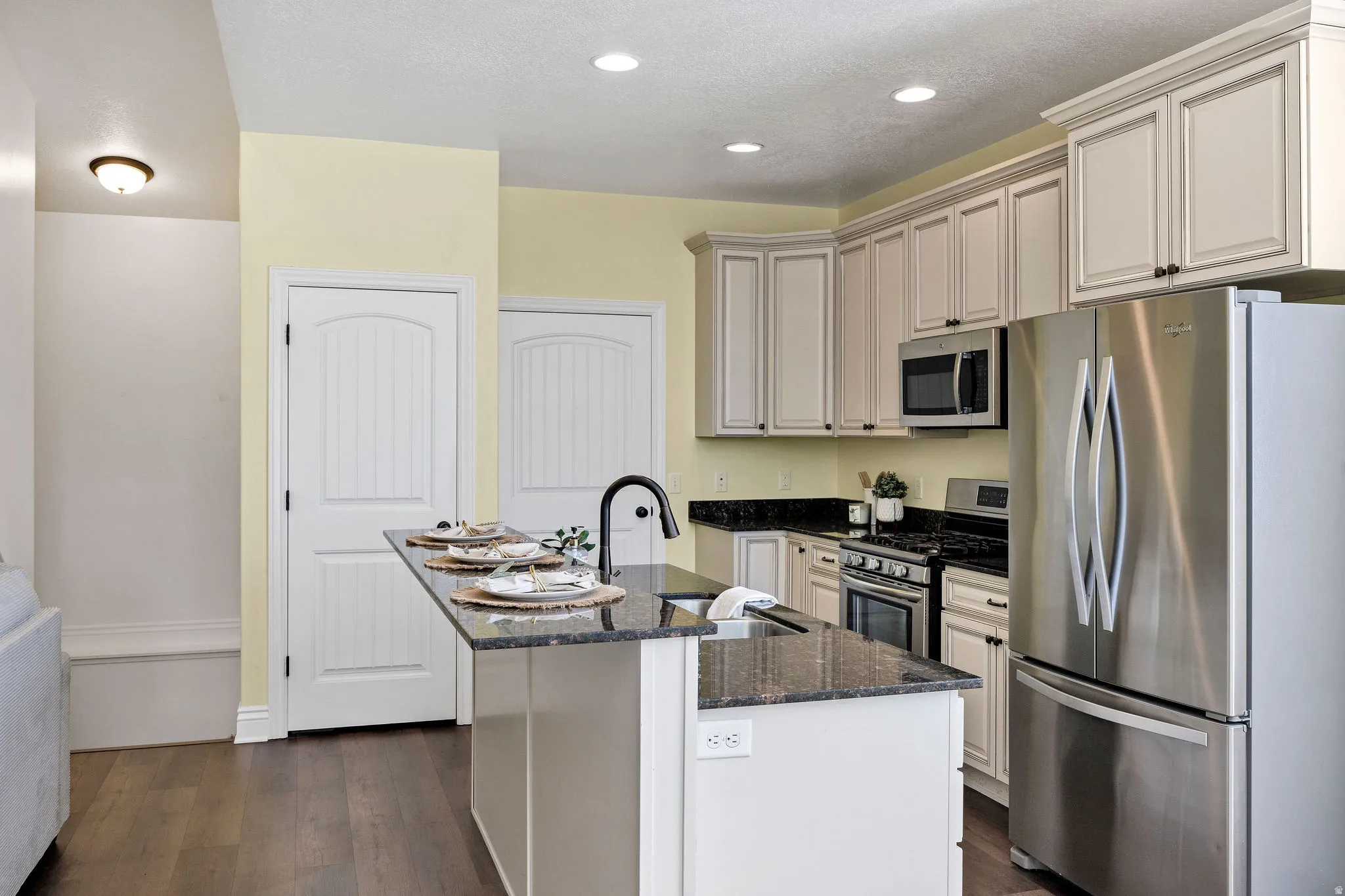 Kitchen with stainless steel appliances, dark stone countertops, an island with sink, dark wood-style floors, and recessed lighting