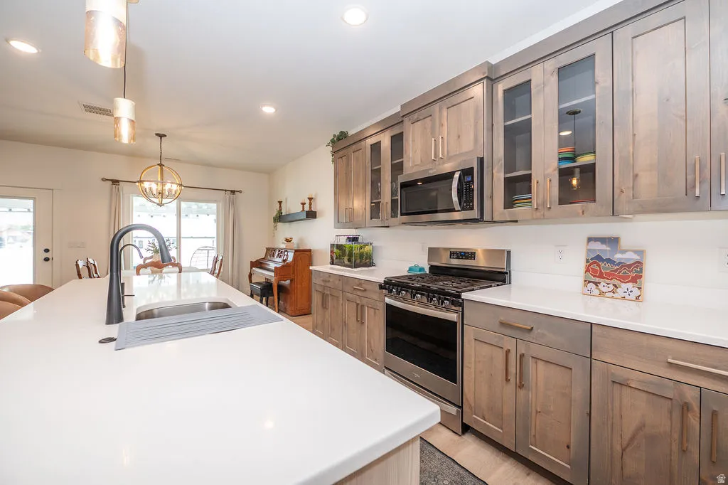 Dual tone kitchen featuring glass insert cabinets, stainless steel appliances, a chandelier, an island with sink, and light stone countertops