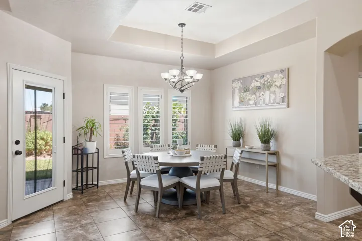 Open dining area featuring tile flooring, a tray ceiling with a chandelier, three windows with plantation shutters, and an exterior door with a full-lite glass panel
