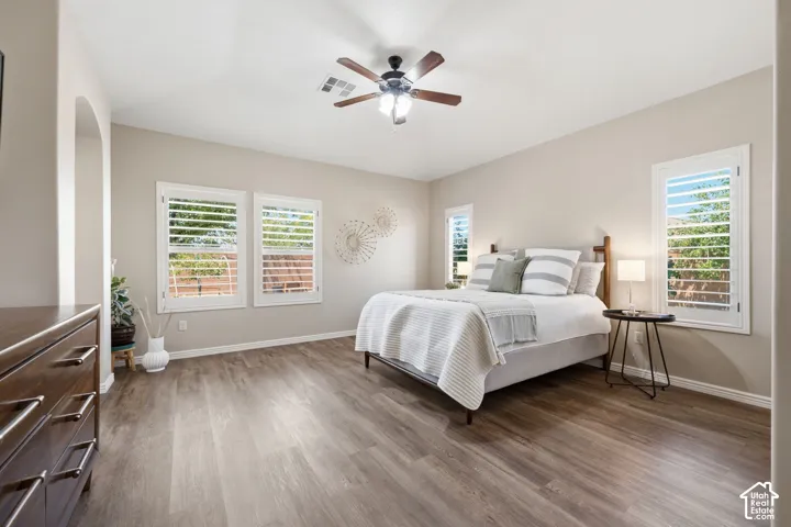 Spacious room featuring wood-finish flooring, multiple windows with white plantation shutters, a ceiling fan with integrated lighting, and an arched doorway