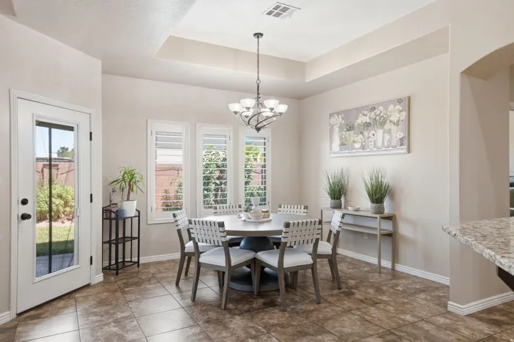 Open dining area featuring tile flooring, a tray ceiling with a chandelier, three windows with plantation shutters, and an exterior door with a full-lite glass panel