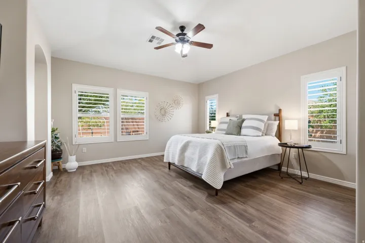 Spacious room featuring wood-finish flooring, multiple windows with white plantation shutters, a ceiling fan with integrated lighting, and an arched doorway