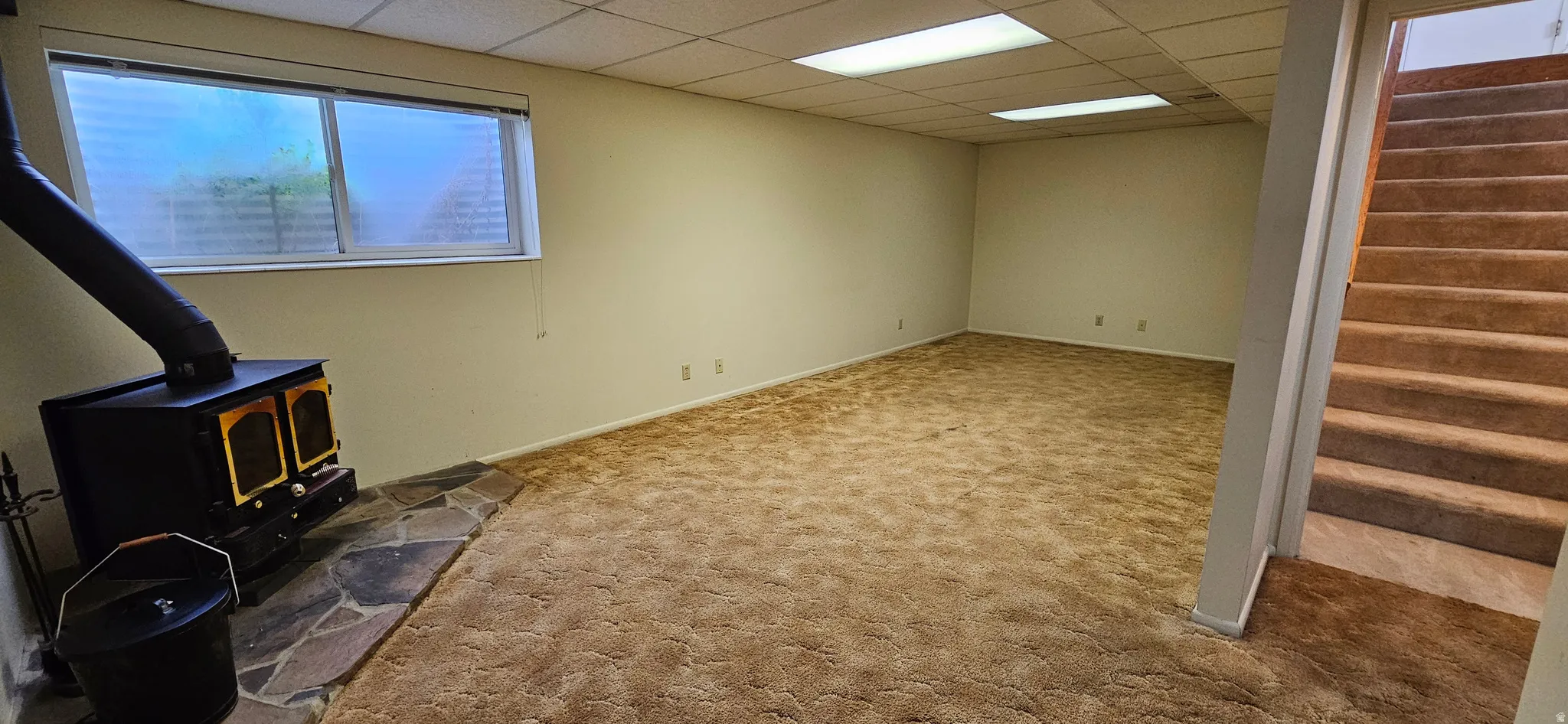 Basement featuring a wood stove, a paneled ceiling, and carpet