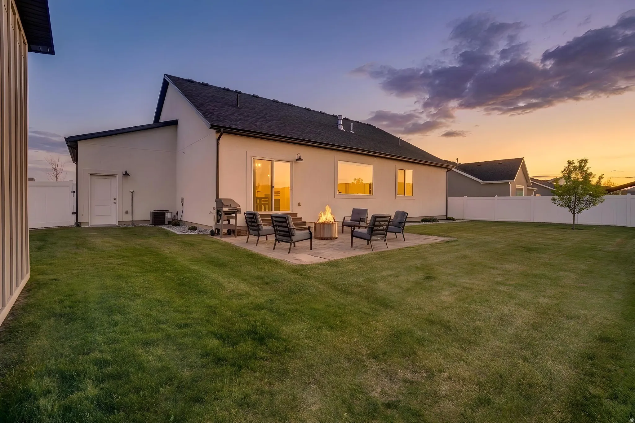 Expansive backyard with a large patio area, featuring a built-in fire pit, a sliding glass door entry, multiple windows, and a white privacy fence