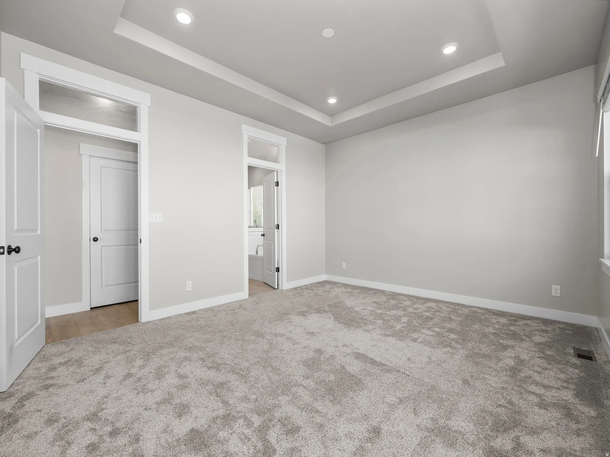 Unfurnished bedroom with light carpet, a raised ceiling, ensuite bath, and recessed lighting