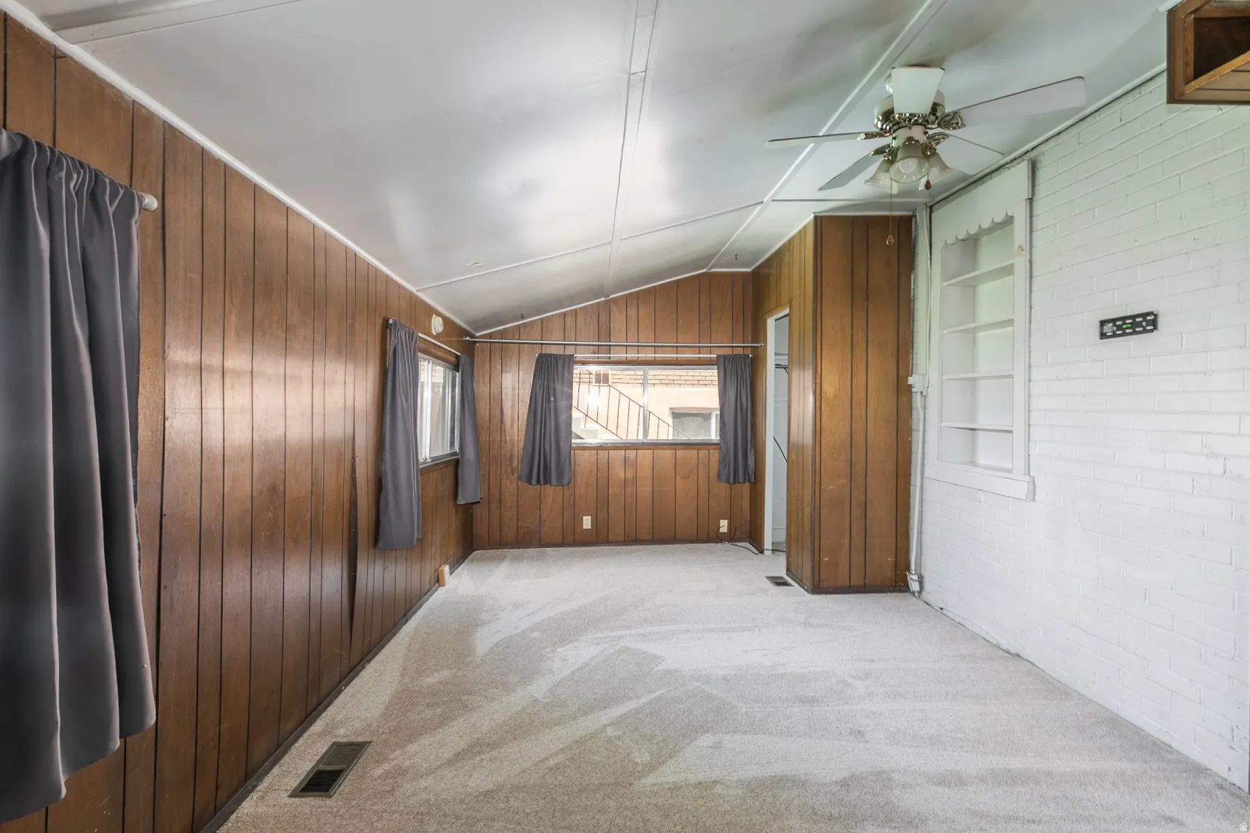 Spare room with light colored carpet, a ceiling fan, vaulted ceiling, wood walls, and brick wall