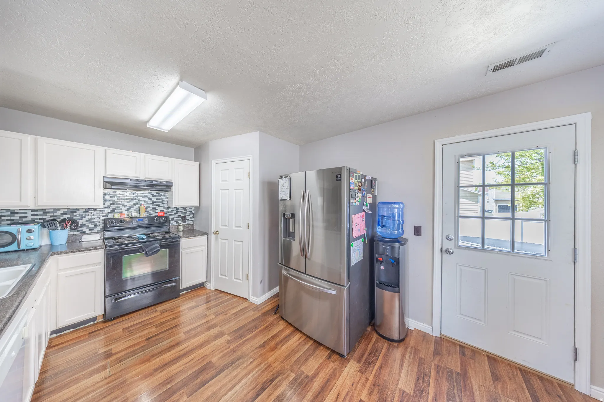 Kitchen with black range with electric cooktop, stainless steel fridge with ice dispenser, white cabinets, decorative backsplash, and light wood finished floors