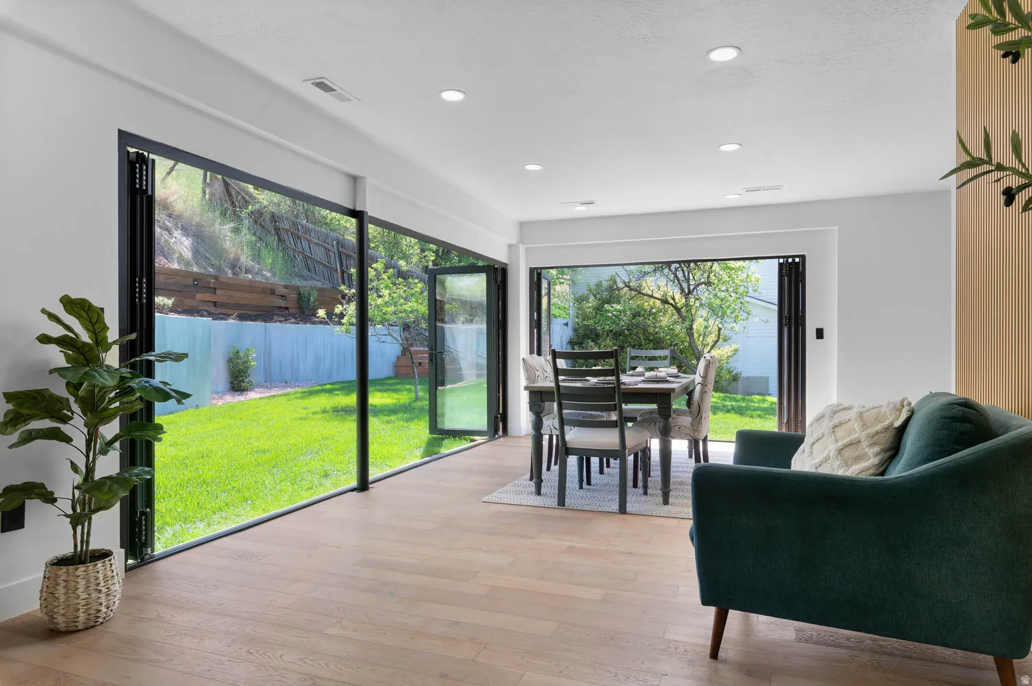 Spacious room featuring floor-to-ceiling bi-fold glass doors, light wood-finish flooring, recessed lighting, and a dining area with a rectangular table and chairs