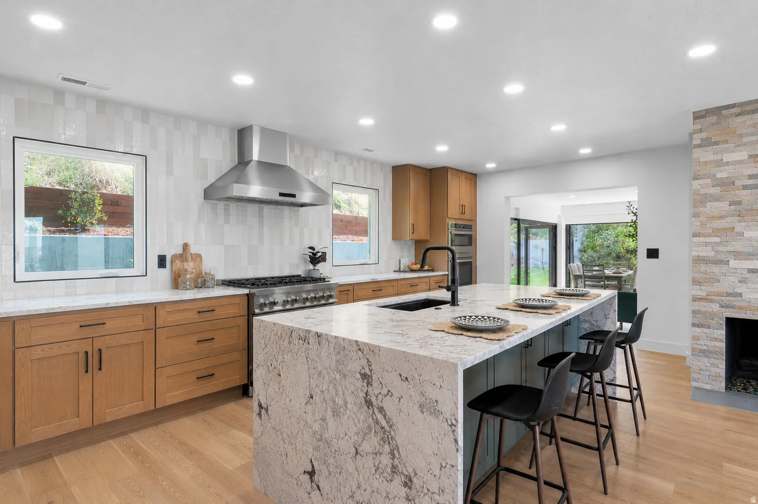 Modern kitchen featuring a large waterfall island with seating, a professional-grade range with a stainless steel vent hood, light wood-finish cabinetry, and a full-height tile backsplash